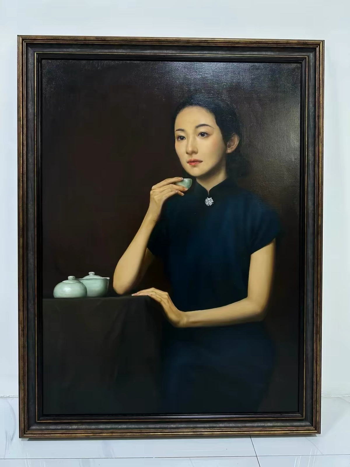 Un gran retrato clásico con marco oscuro de una mujer sentada con un vestido oscuro de cuello alto, sosteniendo una pequeña taza junto a recipientes de cerámica clara.