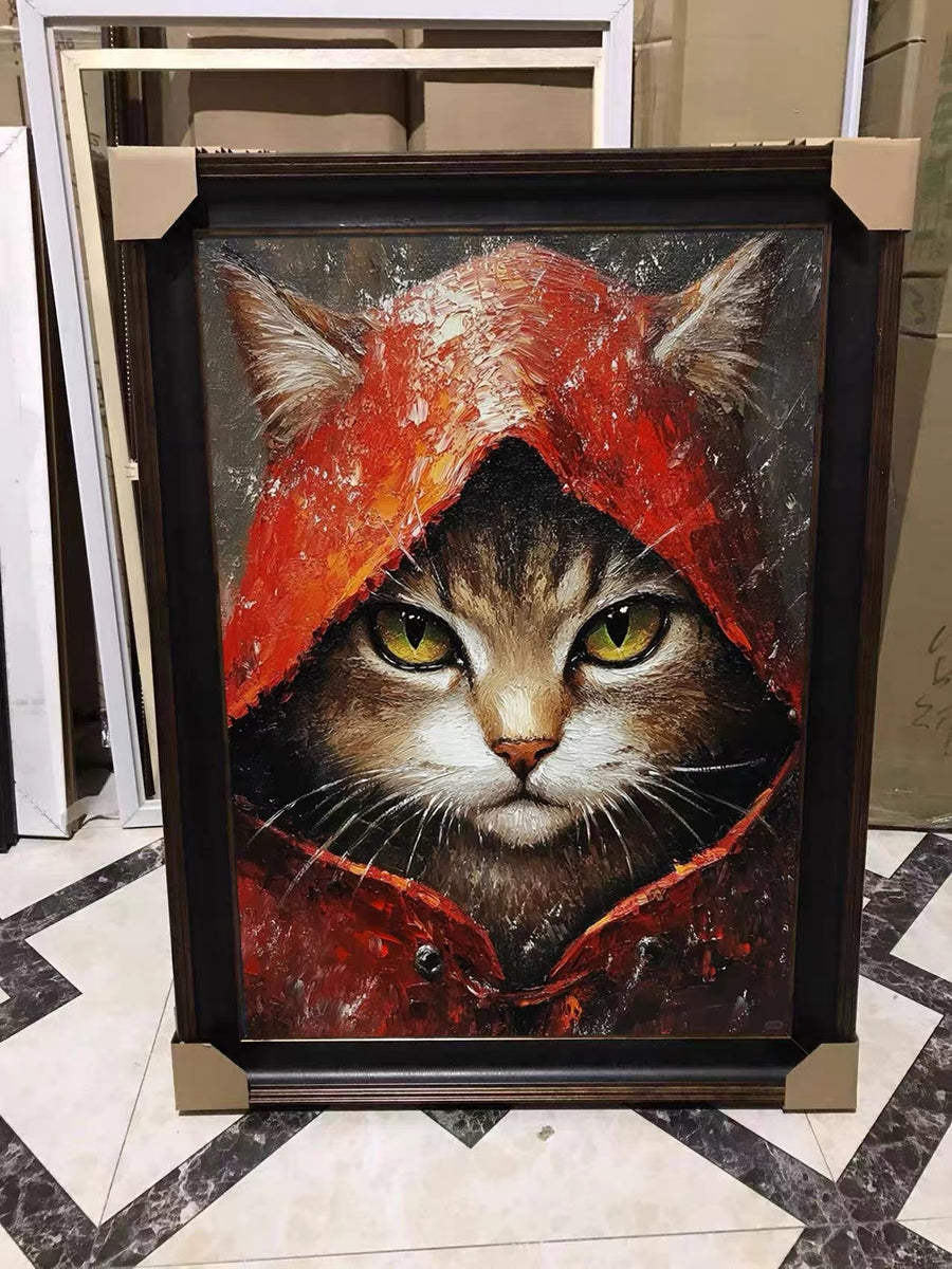 A framed creative portrait of a tabby cat with yellow-green eyes staring forward from inside a red hood.