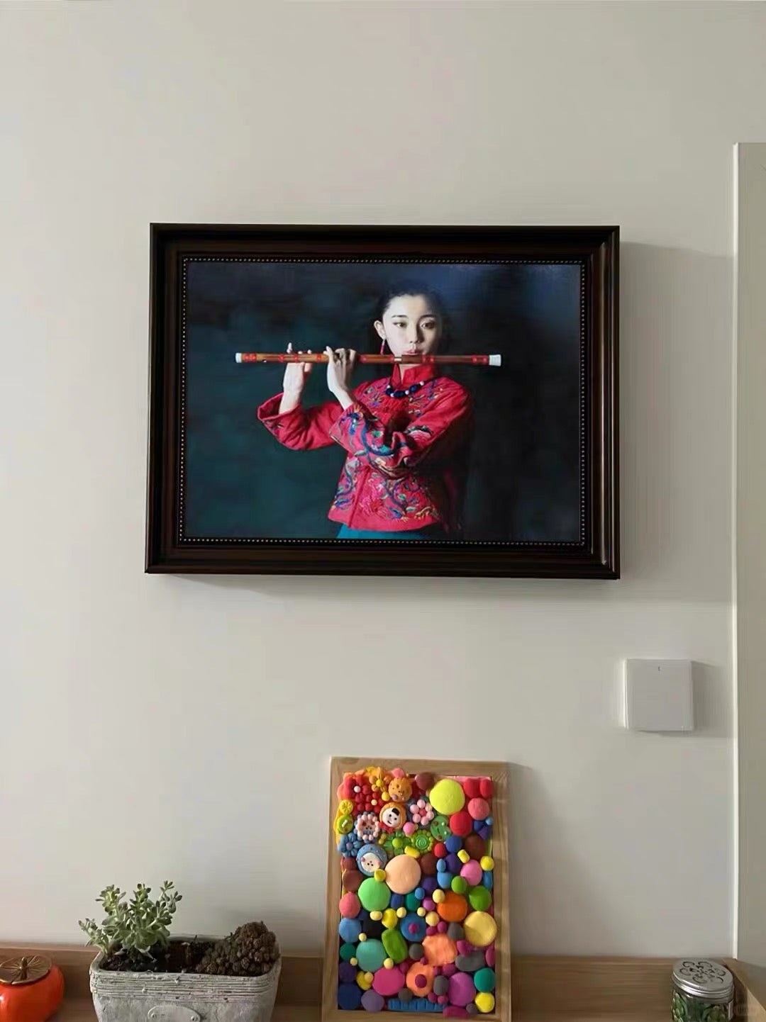 Retrato enmarcado expuesto en una casa, con una mujer vestida de rojo tradicional sosteniendo una flauta sobre un fondo azul oscuro y negro.